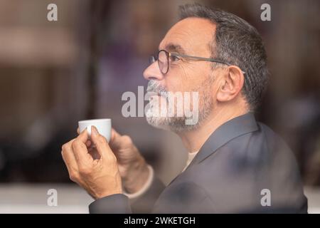 Contemplative mature man with glasses looking out the window, holding a small espresso Stock Photo