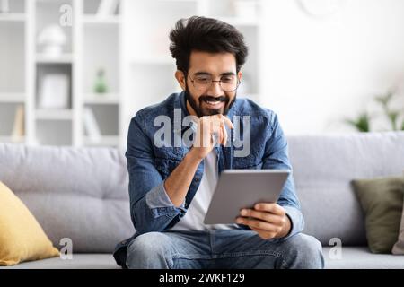 Focused indian man wearing eyeglasses using digital tablet at home Stock Photo