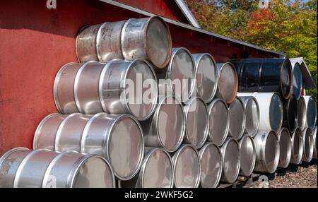 Rows of stacked metal barrels behind a red building, with trees in the background. Stock Photo