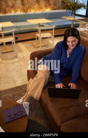Vertical full length portrait of smiling young businesswoman using laptop on couch in coworking office Stock Photo