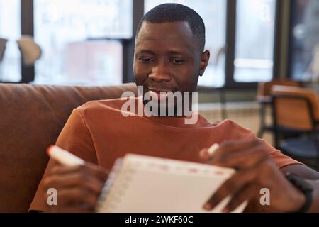 Front view portrait of smiling African American man drawing sketch while sitting on sofa at coffee shop in low lighting copy space Stock Photo