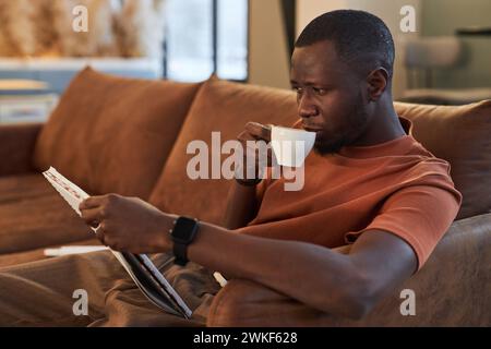 Side view portrait of African American man enjoying cup of coffee while relaxing on couch in cafe and reading document Stock Photo