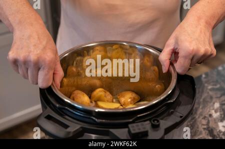 Fingers of a womans hands grasping a metal pot containing cut potatoes while placing it in a modern electric pressure cooker or instant pot. Stock Photo