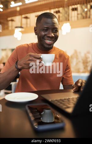 Vertical portrait of smiling young Black man using laptop in coffee shop and enjoying cup of coffee in cozy low light Stock Photo