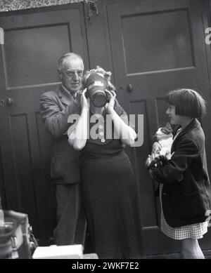 1940s, historical, wartime, a schoolgirl, holding pet cat, watching her grandmother try on a gasmask, England, UK. In the late 1930s, with war with Germany on the horizon, the Britsh government issued gas masks for the public at large, as protection against the dropping of posioned gas bombs, as memories were still strong of the horrors of gas attacks duting WW1. Stock Photo