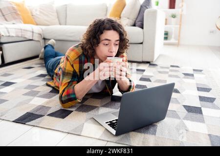 Photo of dreamy funky guy wear plaid shirt lying floor drinking tea watching movie modern gadget indoors room home house Stock Photo