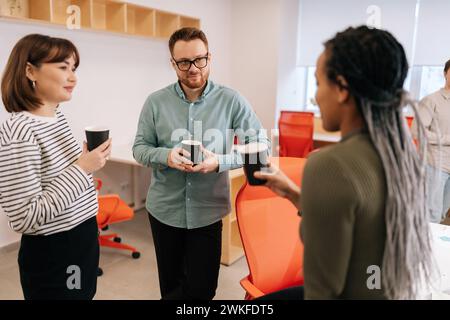 Group of relaxed multiethnic coworkers standing in office, drinking coffee and talking cheerfully in workspace during break. Diverse male and female Stock Photo