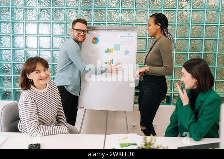 Cheerful two diverse conference speakers drawing on whiteboard during brainstorm meeting with team people in boardroom, give presentation of new Stock Photo