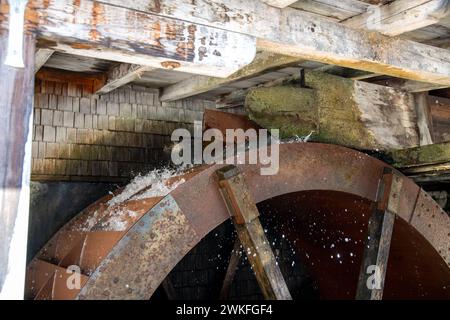 A metal water wheel that is driven by water. The water comes from a wooden frame with an outlet above the waterwheel and runs onto the blades. Stock Photo