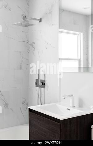 A white bathroom faucet detail with a white sink, modern wood cabinet, and a marble tiled shower. Stock Photo