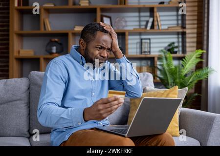 Stressed man holding credit card looking at laptop screen, showing signs of financial trouble and distress. Stock Photo
