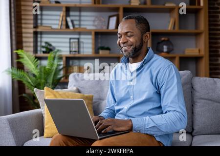 Cheerful African American man working on his laptop while seated on a cozy sofa in a well-decorated living room. Stock Photo