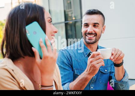 Real handsome man drinking a coffee cup and looking to his girlfriend, she is having a call by phone. Young caucasian couple sitting and relaxing on a coffeeshop terrace. Happy friends taking a break. High quality photo Stock Photo