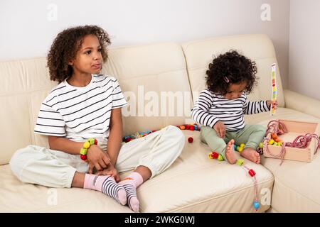 Two African American girls with curly hair are sitting on a couch and playing and occupied with toys Ideal for Stock Photo