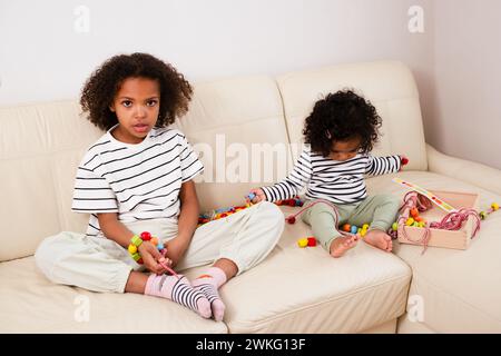 A serious girl in a striped shirt shares a couch with a younger child. Occupied with toys and learning, sibling Stock Photo
