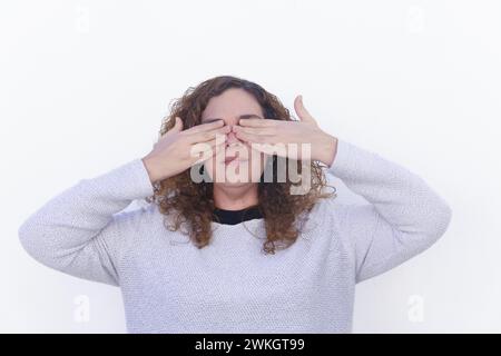 Beautiful blonde curly-haired woman dressed in white, covering her eyes with her hands on a white background and copyspace Stock Photo