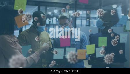 Image of virus cells floating over diverse business people with face masks discussing Stock Photo