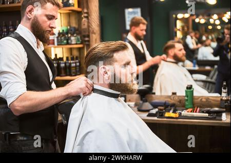 Male barber putting on white cape. Regular client visiting favorite barbershop. Stock Photo