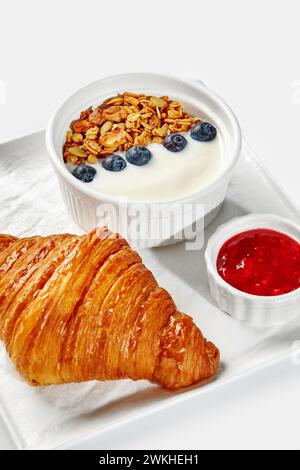 Delicious continental breakfast setup featuring buttery croissant, yogurt topped with granola and blueberries, and side of red berry jam served on whi Stock Photo