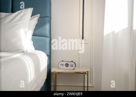 Blue king size bed with crisp white bedding and digital alarm clock on the nightstand under beautiful morning sunlights Stock Photo