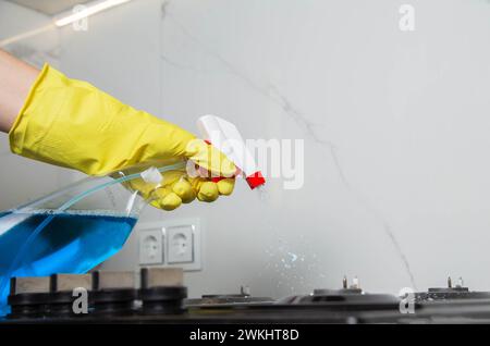 A girl's hand with a cleaning spray sprays on a built-in gas stove. Modern anti-grease, antistatic cleaning product. Background white porcelain tiles Stock Photo