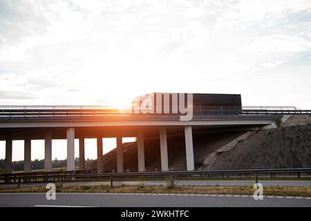 A truck with a curtain-sided semi-trailer transports a load of construction materials in the evening along a bridge against the backdrop of a sunset a Stock Photo