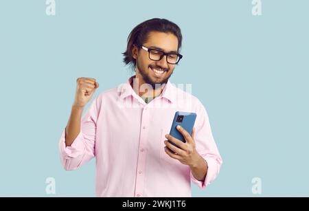 Happy smiling beautiful guy celebrates the victory, holding mobile phone in one hand clenches his fist and raises his other hand up. Stock Photo