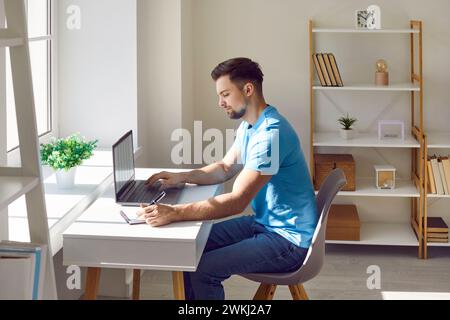 Young man writing notes while using laptop at home Stock Photo