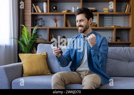 Cheerful bearded man sitting on the sofa in a cozy living room, celebrating a win with his smartphone in hand. Stock Photo