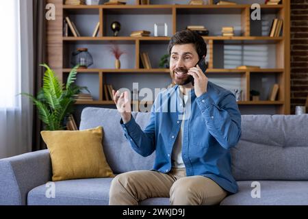 A cheerful man having a pleasant phone conversation while sitting on a couch in a cozy, stylish living room. Stock Photo