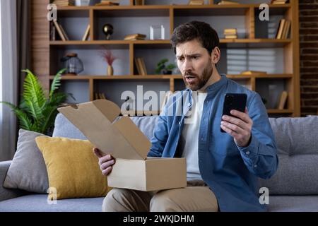 Perplexed bearded man sitting on a sofa, looking at his phone with confusion while holding an open package. Stock Photo