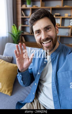 A cheerful, bearded man in casual wear waves at the camera with a warm, welcoming smile in a comfortable home setting. Stock Photo