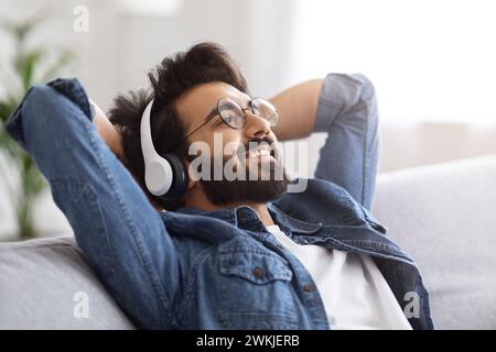 Joyful bearded indian man wearing glasses and wireless headphones reclining on sofa Stock Photo