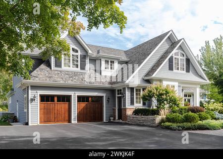 A large grey and white modern farmhouse with two wooden garage doors, and beautiful landscaping. Stock Photo