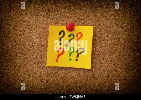 A yellow paper note with colorful question marks on it pinned to a cork board. Close up. Stock Photo
