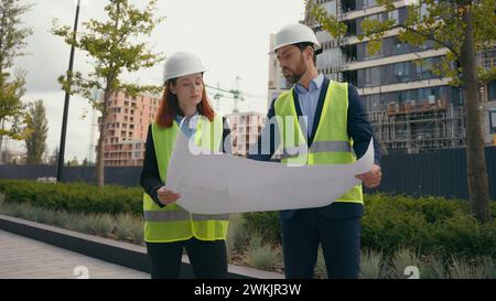 Two people workers builders in safety vests uniform hard hats discuss blueprint document paper plan of unfinished construction site discussion Stock Photo