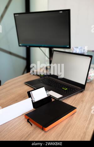 Open laptop with a blank screen, closed notebook with a pen on top, set on a light brown wood table in a cozy workspace setting. Stock Photo