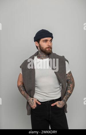 Handsome fashionable hipster man with a beard with tattoos in a fashion white T-shirt with a knitted sweater and hat stands on a white background in t Stock Photo