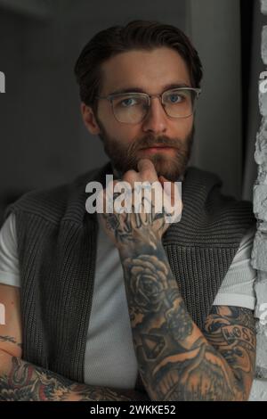 Stylish handsome fashionable hipster man with a hairstyle and beard with a tattoo with fashion glasses looks at the camera in the room Stock Photo