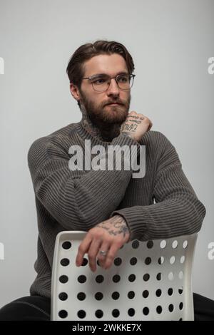 Stylish handsome young hipster man with glasses with hairstyle and beard in vintage stylish knitted sweater sitting on white chairs in studio Stock Photo