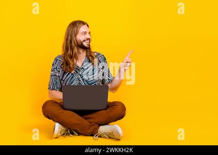 Full length photo of good mood dreamy guy wear print shirt working modern device pointing empty space isolated yellow color background Stock Photo
