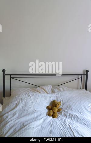 A portrait orientated image of an old teddy bear lying on a bed with an iron frame and white bed linen. Includes space above for text Stock Photo
