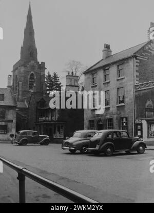 Uppingham Market Place, Rutland, in the 1950s. The image shows the Church of St Peter and St Paul in the background. 1950s cars are parked in the market square. The Grade II listed three story building was once the Doctors Surgery and is now the post office. This photo was taken from the original negative and there maybe blemishes. Stock Photo