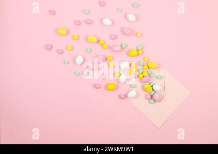 Empty white card in envelope and  scattered chocolate Easter colored candy eggs on pink background. Easter concept. Stock Photo