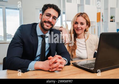 Teamwork of businesspeople sitting on desk and looking at camera. Two startup partners working with a laptop computer at office workplace. Young adult coworkers staring front with confident expression. High quality photo Stock Photo