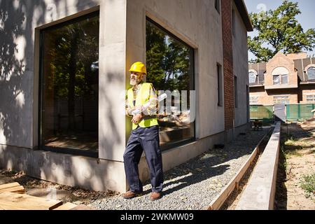 good looking dedicated cottage builder in safety gear with blueprint and posing on construction site Stock Photo