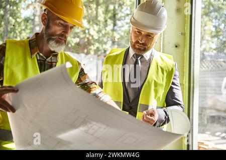 handsome bearded businessman and cottage builder with safety gear working with blueprint on site Stock Photo