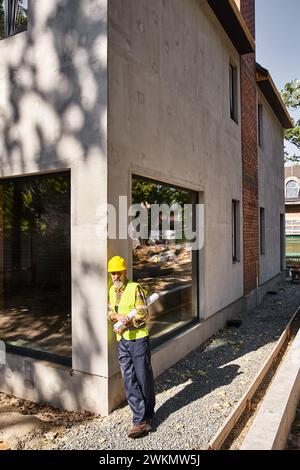 good looking dedicated cottage builder in safety gear with blueprint and posing on construction site Stock Photo