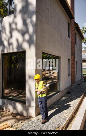 good looking dedicated cottage builder in safety gear with blueprint and posing on construction site Stock Photo