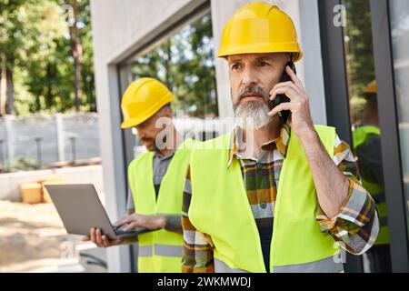 attractive builder with tattoos working on laptop while his colleague talking by phone during work Stock Photo